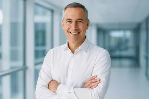 An adult male in a white shirt stands in an office with a smile on his face.