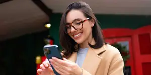 A woman wearing glasses and a coat is smiling and looking at her phone while standing in front of a red door.