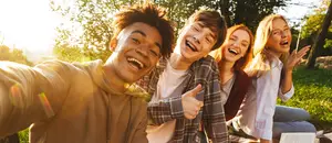Four young people, including two women and two men, are sitting on the grass and posing for a selfie, smiling and laughing together