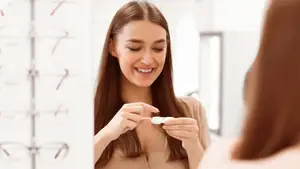 A smiling woman holding a contact lens in front of a mirror