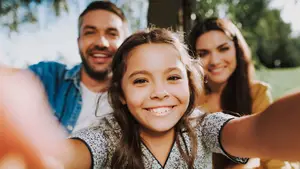 A smiling young girl and her parents are taking a selfie in a park with trees in the background.