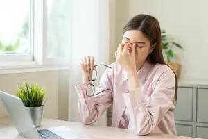 A woman wearing glasses is sitting at a desk with her hands covering her eyes, perhaps looking at her laptop.
