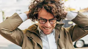 A smiling man with curly hair wearing glasses and a jacket is standing on a street and posing for a photograph with his hands on his head.