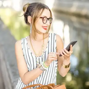 A smiling woman with glasses and jewelry looking at her phone on a bridge