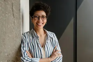 An adult woman with glasses and a striped shirt smiles while leaning against a wall with her arms crossed