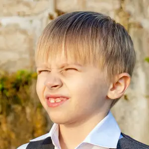 A boy with a smiley face and a slight frown is wearing a white shirt and a black vest, standing in front of a stone wall with some plants behind him.