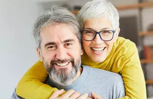 An elderly woman with glasses hugging a man with a beard and gray hair in a room