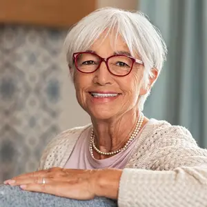 Portrait of a smiling elderly woman with white hair, wearing glasses, a pearl necklace, and a beige sweater, sitting in a cozy room with a blue curtain and a wooden shelf in the background.