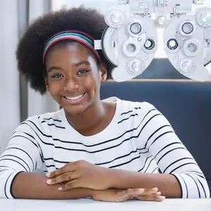 A young girl with afro hair smiling while sitting in front of an optometrist's eye exam equipment.