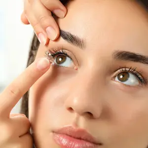 A woman is touching her eye with a finger and a lens on her finger. She has long eyelashes and is wearing makeup.