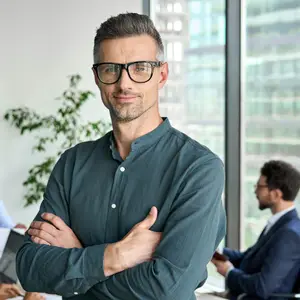 A businessman wearing glasses is standing with his arms crossed in an office with another man sitting behind him.