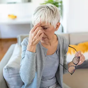 A lady with glasses, sitting on a couch, and looking at something with her eyes closed.