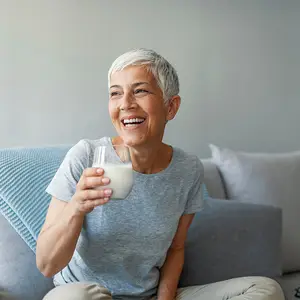 An older woman is sitting on a couch, smiling and holding a glass of milk in her right hand.
