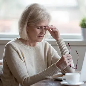 A woman in a beige sweater sitting at a table with a laptop and a cup of coffee looks stressed.