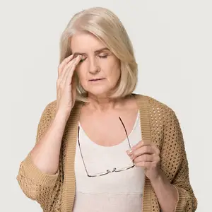 An elderly woman wearing glasses and a beige cardigan is holding her forehead with her left hand, possibly feeling tired or experiencing a headache. She is looking down with her eyes closed, possibly in a room. She is holding a pair of glasses with her right hand, which has a silver frame. She is probably trying to relieve her headache by resting her head on her hand.