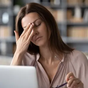A woman sitting at a desk with her hand on her forehead, wearing glasses and a pink blouse.