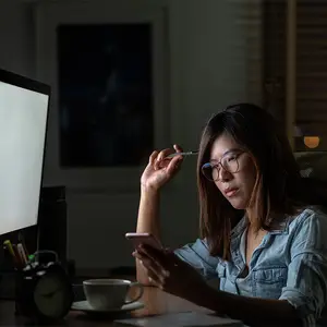 A woman is sitting at a desk with a computer monitor, holding a phone and pen, and looking at her phone in a dimly lit room.