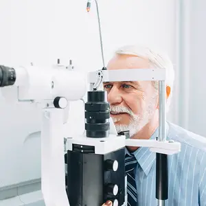 An older man is smiling while examining his eyes with an ophthalmoscope in a medical room.