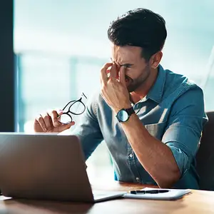 A man is sitting at a desk and rubbing his eyes with his hand, he is wearing glasses and a watch and there is a laptop and a notepad in front of him.
