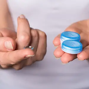 A person's hand holding a blue lens with a white lens and a blue bottle with a white lens inside on a white background.