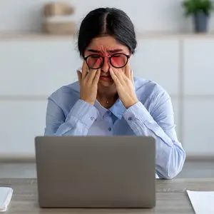 Woman sitting at a desk, holding her head and rubbing her eyes in pain.