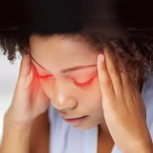A close-up of a woman with curly hair holding her head with both hands and looking down with red eyes.