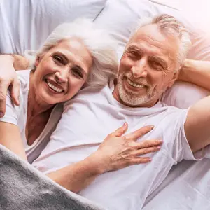 An elderly couple smiles and poses for a photo in their bedroom