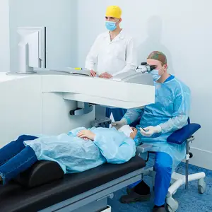Three people in a room, one is lying on a table while two others are wearing blue gowns and face masks and one of them is using a microscope