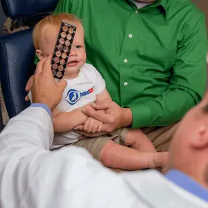 A young child is having their eyes examined by an adult male doctor while being held by a man in a green shirt.