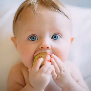A baby with blue eyes is holding a wooden toy in its mouth while lying on a white surface.