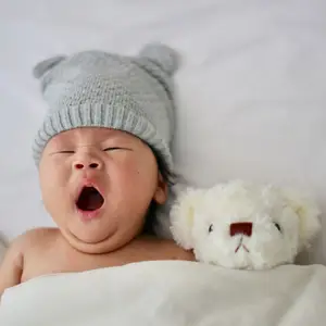 A baby lying on a bed, yawning, and wearing a gray knitted hat is next to a white teddy bear