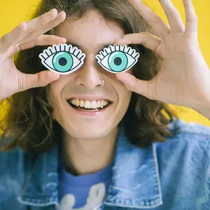 A smiling person wearing denim jacket and holding up glasses with green eyes and white circles