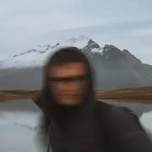A person wearing a black hoodie jacket stands in front of a body of water with a mountain covered in snow in the background. The person's face is obscured by the hood and the image is slightly blurry.