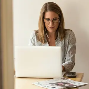 Woman sitting at a table with a laptop, headphones, and a newspaper on the table in front of her.