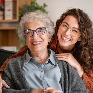 A woman with gray hair wearing eyeglasses sitting on a couch and a young woman with curly hair wearing eyeglasses leaning on her shoulder and smiling