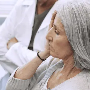 A woman in a white sweater sitting in a medical office looking concerned while touching her face