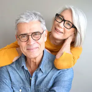 A smiling elderly couple posing for a photograph, with the woman resting her hand on the man's shoulder