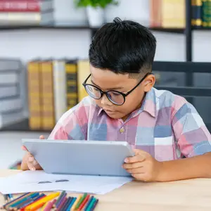 A young boy wearing glasses is sitting on a chair and seems to be doing something on his tablet