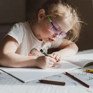 A young girl with glasses is drawing on a piece of paper with a crayon