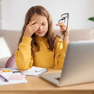 young girl sitting at a desk with a laptop holding glasses and looking tired