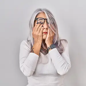 An older woman in a white long sleeve shirt and glasses covering her eyes with her hands