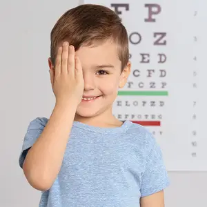 A smiling boy with brown hair wearing a blue t-shirt, touching his eye with his hand while standing in front of an eye chart.