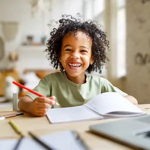 A young child is smiling while sitting at a desk with a notebook and pencil.