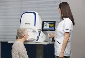 An elderly woman is sitting in front of a Nikon device in a medical room, a nurse is standing beside her, and the nurse is holding a remote control.