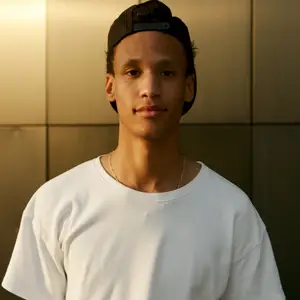 A man with a black cap and white t-shirt standing in front of a beige wall