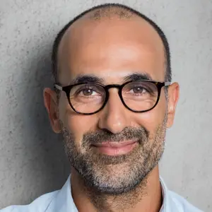An adult man with a beard and glasses wearing a light blue shirt is smiling for a photo with a textured wall in the background