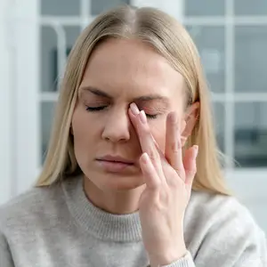 A blonde-haired woman with her eyes closed, gently touching her forehead with her fingers, possibly in a room with a white wall and glass window behind her.
