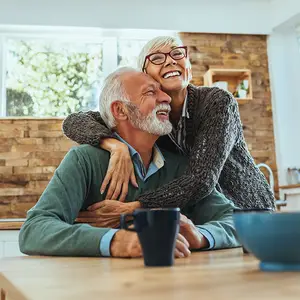 Elderly couple sitting at a table with cups and bowls, smiling and embracing