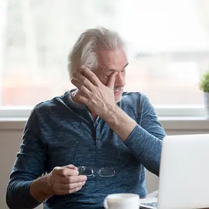 An elderly man is sitting in front of a laptop, holding a pair of glasses and touching his forehead with his left hand.