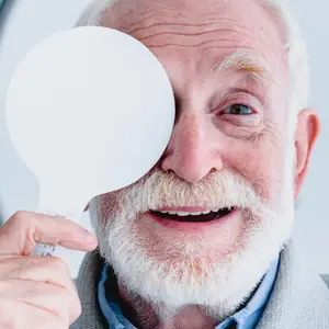 An elderly man wearing a sweater is smiling and holding a white magnifying glass in front of his left eye.